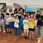 興仁國小學生與師長在兒童音樂劇始業式中合影，手持玩聚學習與教育公益標語