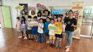 興仁國小學生與師長在兒童音樂劇始業式中合影，手持玩聚學習與教育公益標語