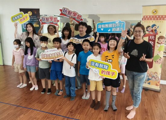 興仁國小學生與師長在兒童音樂劇始業式中合影,手持玩聚學習與教育公益標語